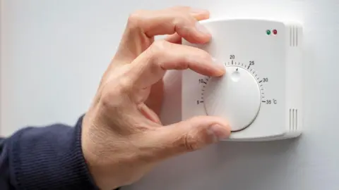 A mans hand adjusts a white thermostat on a white wall. The mans arm is covered by a navy sweatshirt. 