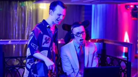A screenshot from Kirkmoore. A man in a jazzy shirt stands behind Keron's character who is sitting in front of his a computer screen and wears a headseat and a suit. They are are in a bar or club.