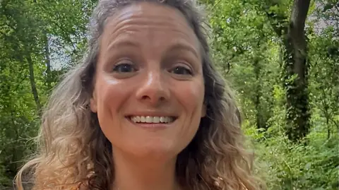 Zoe smiles at the camera in a selfie. She has light brown, curly hair and there are trees and woodland visible behind her. 
