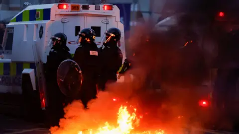 Reuters armed police officers wearing black uniforms stand behind white police. A flame rose into the air behind him.