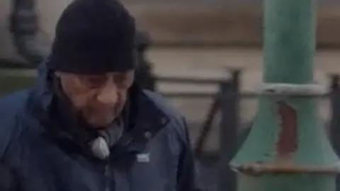 An elderly man using a walking stick and wearing a black hat and long coat walking towards court with help from someone who is blurred out.