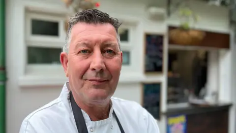 A man with grey hair, wearing a white chef shirt and a dark blue apron, is looking into the camera. He's standing in front of the cafe he owns, and there are two black boards on the white building wall behind him and a hatch for serving.