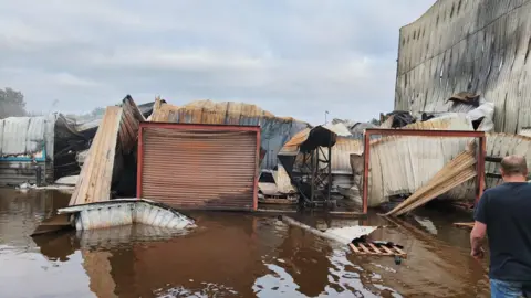 Essex Packaging Supplies The remains of a building that has been flooded following a fire. Scraps of metal are collapsed around what were a pair of garage doors. Brown water is on the ground. The back of a man wearing a blue T-shirt is at the right of the frame. 