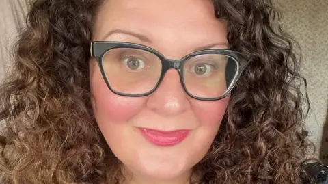 Kathryn Stonehouse A woman with glasses and curly dark hair, she smiles at the camera in a selfie-style photograph.