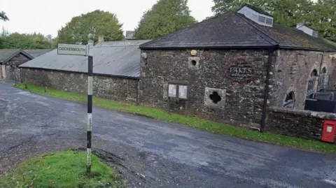 Google The Lakes Distillery. The name of the businesses has been painted onto a stone building. A black and white signpost points to Cockermouth.