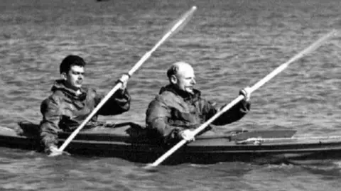 National Museum of the Royal Navy A black and white photograph of two men paddling in a two-man canoe in the sea.
