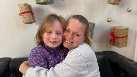 Charlie Jones/BBC Maya (left) wearing a purple top cuddles her mum Marta (right) who wears a grey jumper