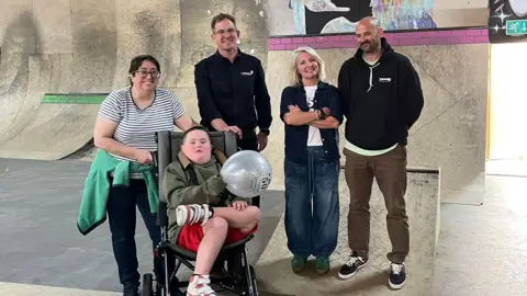 Exist Skatepark A woman holding a wheelchair with Ollie sat in it. Behind him is three people which include Dr Rob Head and the two owners of Exist Skatepark. You can see a skate ramp behind them.