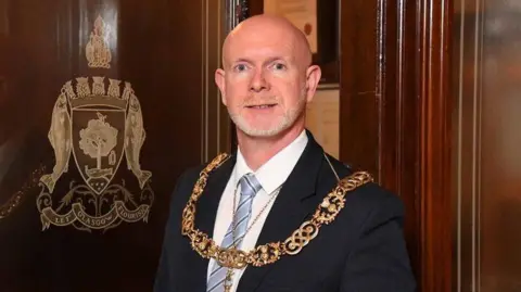 A bald man wearing a large gold chain, black suit jacket, white shirt and blue striped tie stands next to a wooden door which has the gold-embossed Glasgow coat of arms on it