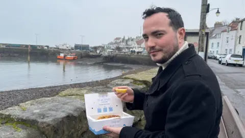James Muirhead, who is wearing a dark coat, sits by a harbour on a grey day, holding a takeaway box of fish and chips and a lemon wedge. Fishing boats rest in the background near a stone pier, with pastel-coloured houses lining the waterfront.