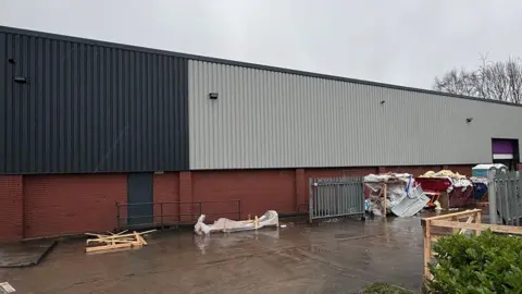 The long warehouse is lined with dark and grey metal at the top, and red-brick at the bottom. At one end is a doorway with a pinky purple jamb. In front of the building is a wide concrete space with assorted bits and pieces scattered around - some broken wooden pallets, some wooden things wrapped in plastic, some fencing and a skip of rubbish. Near the door is a portable toilet.