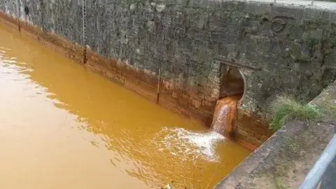 A culvert/drain showing stained orangey-brown water pouring into Queens Dock. There is a tide mark from contaminated water can be seen on the harbour's internal wall.