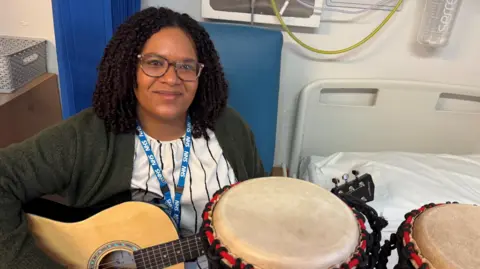 Abbie Jean-Baptiste, a woman with dark hair and glasses, is seated holding a guitar next to some drums. An empty hospital bed is next to her.