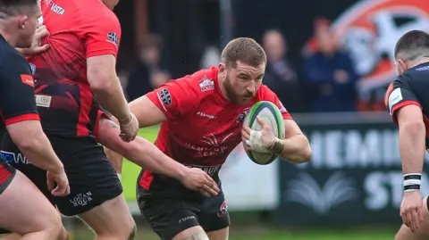 Brian Tempest A bearded rugby player wears a red shirt with black shorts, prepares for the tackle 