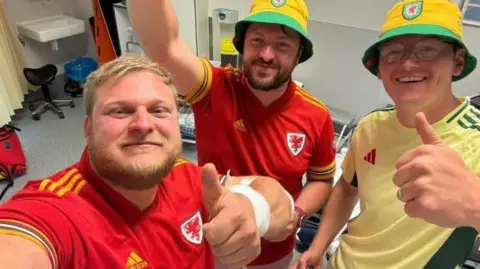 Sam Rees, left, pictured holding his thumb up taking a selfie. He has blonde hair and a blonde beard. Two of his friends can be seen beside him, one with his arm in the air (middle) and another with his thumb up (right) both smile at the camera. 