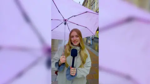 Girl holding pink umbrella and black microphone. She has long blonde hair and is wearing a beige coat.