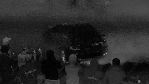 Black and white CCTV camera image showing small crowd watching a car do doughnuts