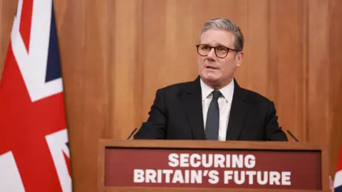 Prime Minister Sir Keir Starmer, wearing a dark suit and tie, speaks behind a lectern displaying the phrase Securing Britain's Future during a press conference on the Immigration White Paper in the Downing Street Briefing Room in London.