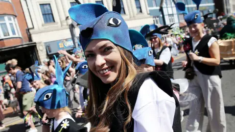 Paul Norris A woman smiles at the camera during the festival parade wearing a costume comprising of a blue glittery hat with ears and eyes. She is wearing a white t-shirt and black tank top with white and yellow daisies. There are other people in the parade wearing the same outfit. Crowds line the parade route in bright sunshine.