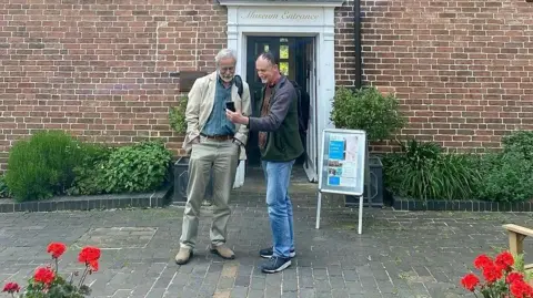 Two men stood looking at a phone in front of a brick building, there are red flowers and greenery outside the building