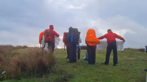Plymouth College A group of teenagers stood holding a map as they try and navigate around on the moors. They are all wearing red rain jackets and big rucksacks on their backs.