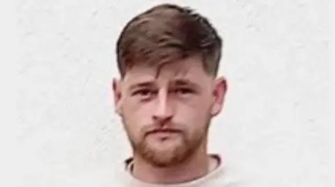 Alex stood against a white background. He is wearing a white jumper and is looking straight ahead. He has brown hair and a short brown beard. 