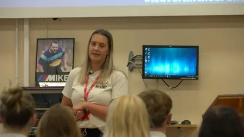 BBC A woman in a white T-shirt stands in front of 10th grade students in a school corridor. Behind him is a photo of his brother, who was stabbed to death.