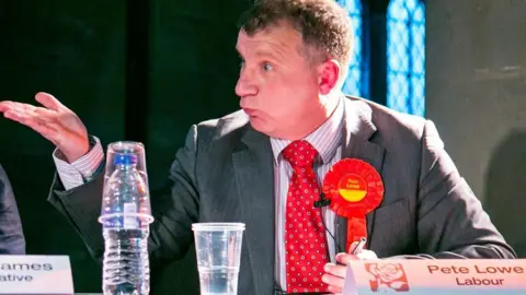 Pete Lowe Councillor Pete Lowe takes part in a political debate wearing a red Labour election rosette. There is a name card and a bottle of water and a plastic cup in front of him. He is holding a hand up as he speaks to make a point.