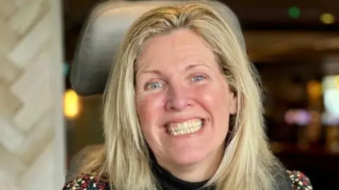 Nina Parry, who has straight blonde hair and is smiling widely at the camera. She is sitting in a wheelchair, with its headrest visible behind her. She appears to be in a pub or a restaurant, although the background is blurred.