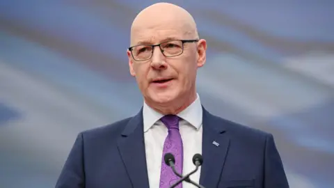 John Swinney, who is bald with glasses, speaks into a microphone. He is wearing a business suit and purple shirt 