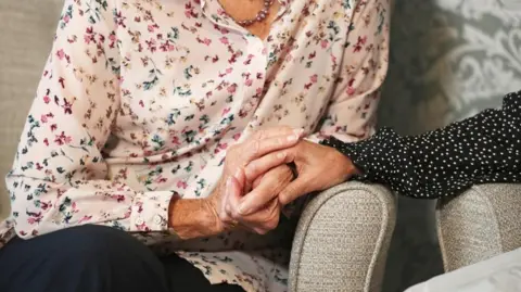An elderly lady wearing a light pink blouse holds the hand of a woman wearing a black shirt with small white dots. They are sat on grey, fabric cushioned seats. 