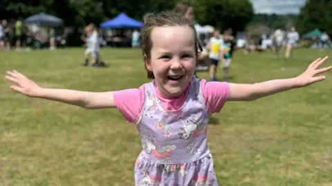 Elizabeth A young girl with a lilac unicorn dress looks at a camera smiling with her hands outstretched. She is in a field. It is a sunny day 