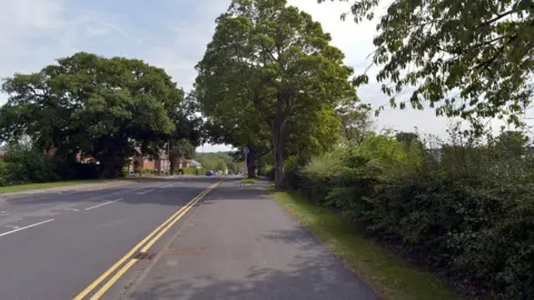 habiloid/Geograph A stretch of A road with double yellow lines, lined with trees and pavement.