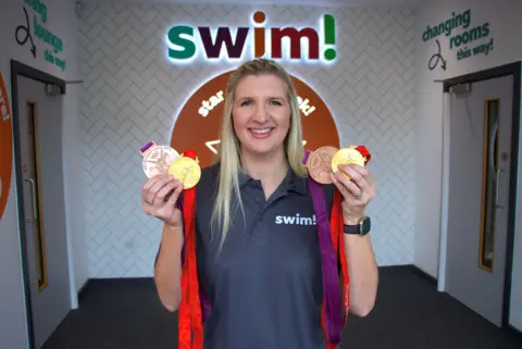 Becky Adlington holds up her medals. She is wearing a navy t-shirt with the word 'swim!' written in light blue writing. She is standing in front of signage that says Swim! in different coloured letters.  