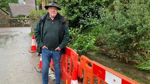 BBC Andy Munro wearing a hat and coat by barriers, near the stream that burst its banks