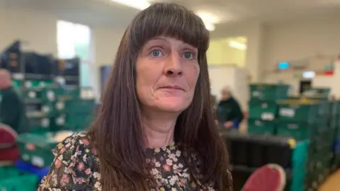 Samantha Price standing amongst food parcels at Rhymney Valley Foodbank looking straight at the camera. Her long dark hair is untied and she is wearing a green dress which is printed with many flowers in different shades of pink, white and red.