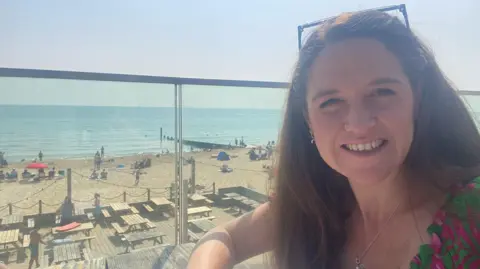 Contributed A woman in a selfie image on a balcony overlooking a beach on a sunny day. She has long dark hair with sunglasses resting on top of her head. She is wearing a green and pink top.