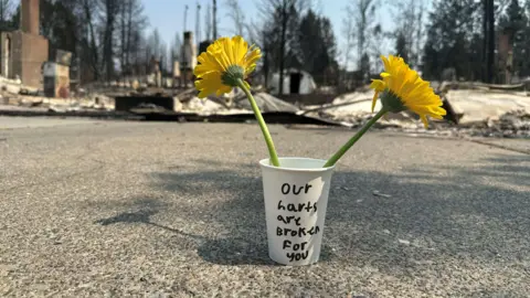 Flowers in a cup with a message to survivors and wreckage in the background. "Our harts are broken for you," it reads