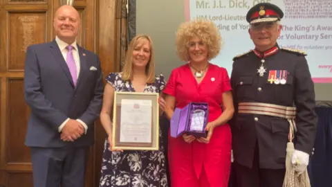 Chris Blacksell, Deputy-lieutenant for East Riding of Yorkshire, Vickie Hillier and Sam Barlow, Fitmums & Friends and Mr James Dick OBE, Lord Lieutenant of East Riding of Yorkshire stand in a line. Vickie Hillier is holding a framed certificate and Sam Barlow is holding a blue box.