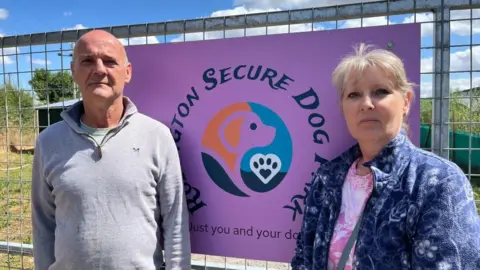 Ellen Knight/BBC Stuart and Sarah photographed in front of a sign for their business. Stuart is wearing a grey quarter zip jumper, and Sarah is wearing a blue floral fleece over a pink t-shirt. The sign behind them is purple, and reads "Rodington Secure Dog Park" in dark blue letters. It has a logo with the silhouette of a dog against a blue and orange background. 