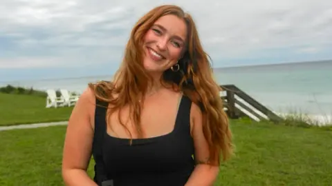 Lizzy has long reddish hair and is wearing a black top whilst standing on a grassy cliff near the beach. The sky is cloudy and wooden steps to the beach and white chairs are visible behind her.