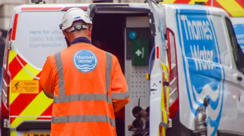 Getty Images A man in all orange high vis and a white hard hat stood behind a Thames Water van.