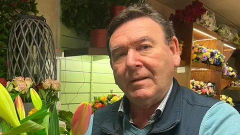 Michael O'Neill is holding a large bouquet of flowers. He is wearing a navy gilet and green fleece, dark hair and is staring in to the camera