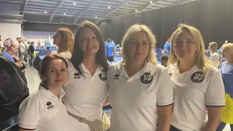 Three women wearing white T-shirts in front of a pickleball court 