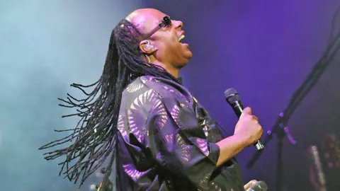 BBC Stevie Wonder holding a microphone and singing with his head lifted up. He is wearing black sunglasses and had long dreadlocks reaching down below his shoulders. He is wearing a purple short sleeve shirt which fades to black, with circles containing a sun pattern on it. 