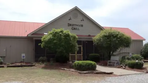 CBS The exterior of the Driftwood restaurant. It is a low building with light brown siding and a reed roof. The name is in capped letters at the top of the building, which comes to a peak. There are trees and a bench outside, along a walkway that leads to a double screen door.