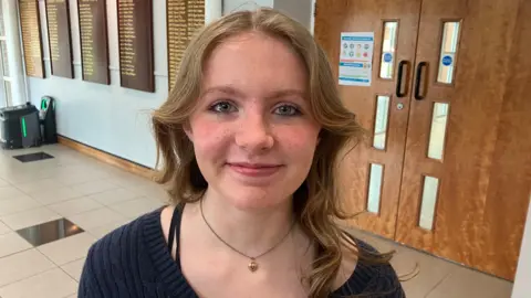 Kaitlyn is a teenage girl with long fair hair and blue eyes. She is wearing a dark blue v-neck top and is looking at the camera. 