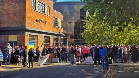 Bradley Tai/Curve Fans stand outside a town centre pub during a football match day. They stand talking to each other many with drinks in hand.