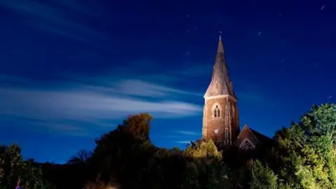 A church spire is illuminated against a dark blue sky with soft clouds and stars. green bushes are in the foreground.