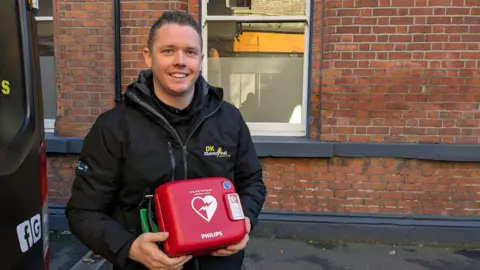 BBC Dale Lee, a smiling man with short hair, holding a defibrillator with the words HEARTSTART/PHILLIPS on it. He is standing on the street, at the back of a van